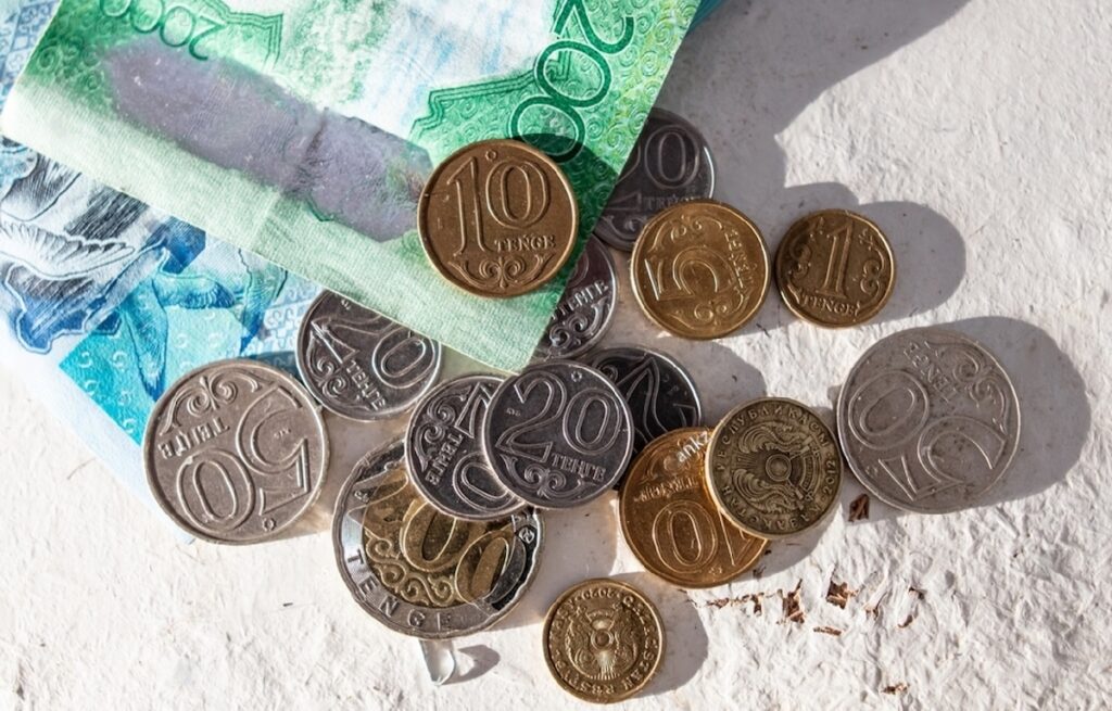 Монеты тенге лежат рядом с купюрами тенге на столе с тенью от солнца