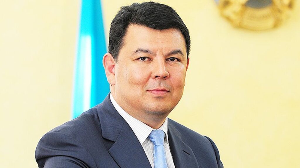 Канат Бозумбаев стоит на фоне флага и герба Казахстана