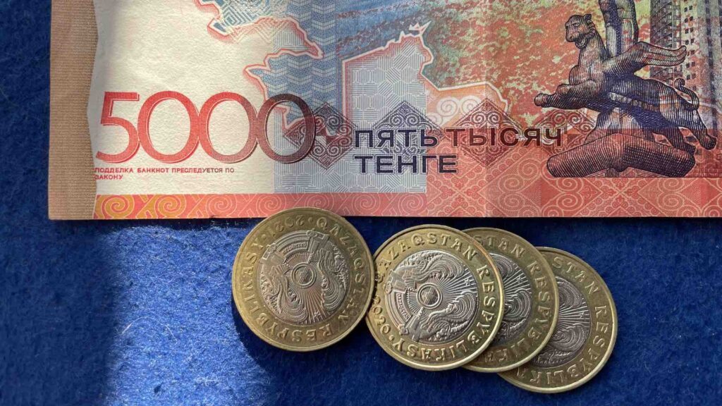 Купюра в 5000 тенге и монеты 100 тенге лежат на синем фоне