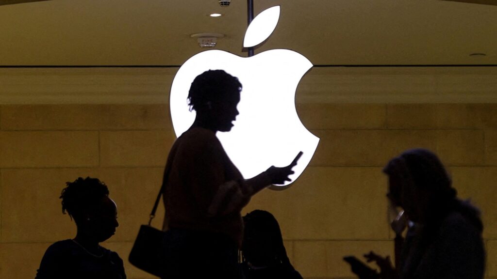 Женщина пользуется мобильным устройством iPhone, проходя мимо освещенного логотипа Apple в магазине Apple в Grand Central Terminal в Нью-Йорке, США.