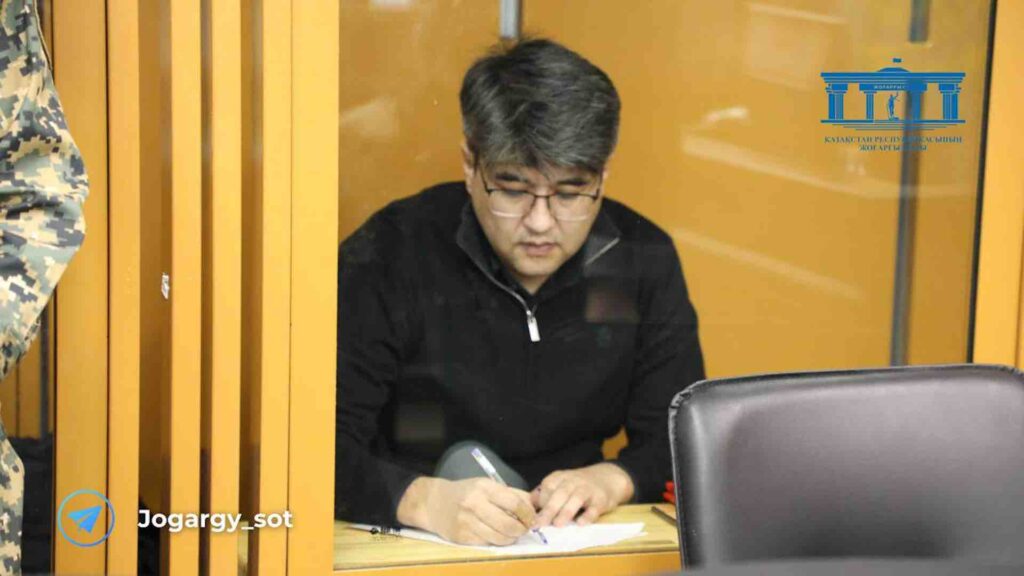 Куандын Бишимбаев пишет ручкой на листке бумаги на скамье подсудимых