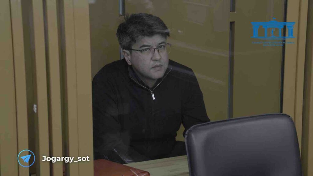 Куандык Бишимбаев сидит на скамье подсудимых и смотрит на камеру
