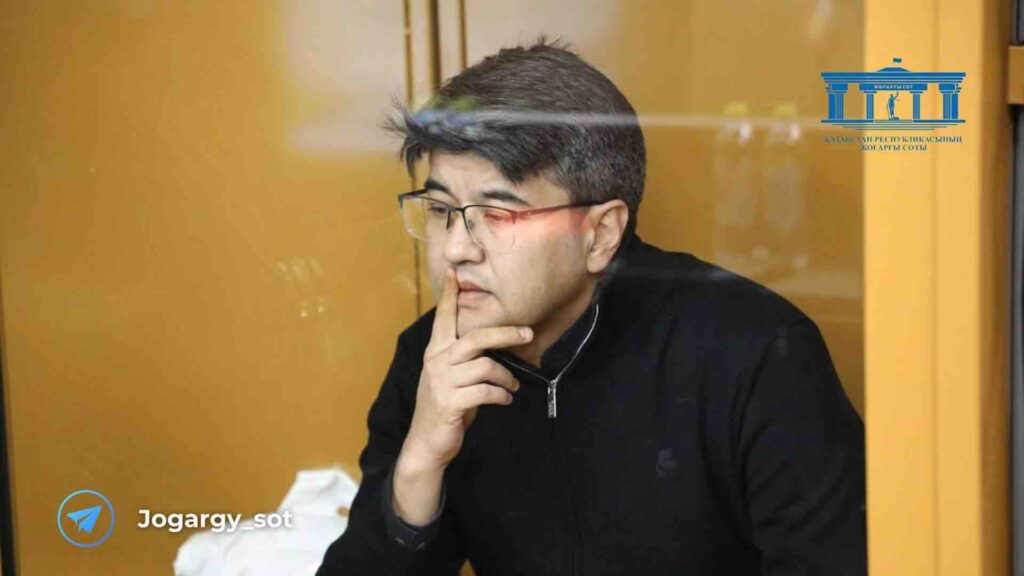 Куандык Бишимбаев в очках и с пальцем на лице сидит на скамье подсудимых