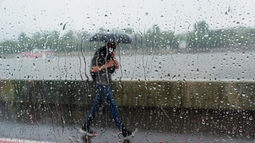 Мужчина идет с зонтом под дождем за стеклом