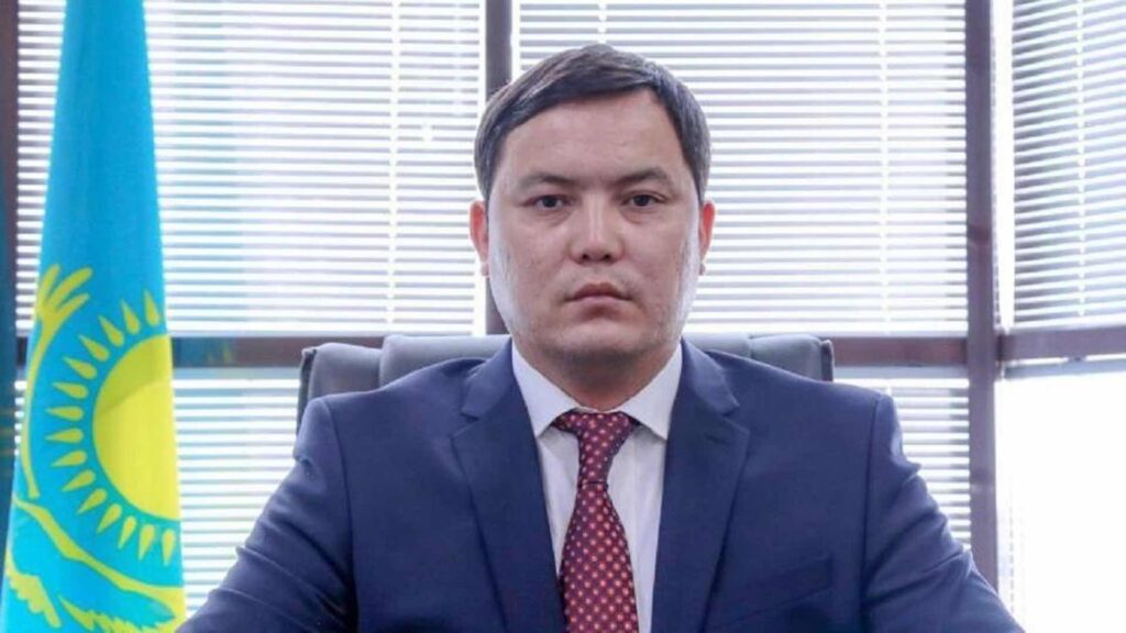 Ринат Ибрагимов в костюме сидит за столом рядом с флагом Казахстана